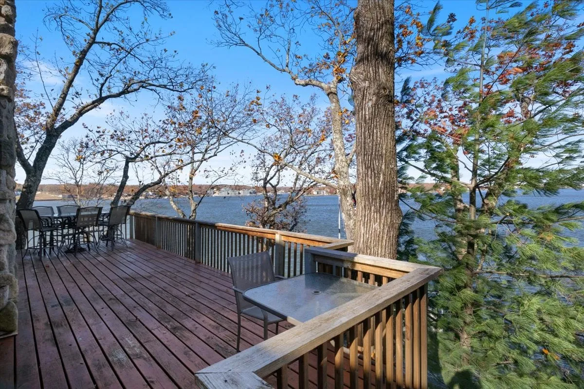 A serene exterior deck outfitted with comfortable seating and dining furniture, overlooking the shimmering lake and surrounded by trees. The setting is ideal for outdoor meals, morning coffee, or unwinding while soaking in the lake views, reinforcing the indoor-outdoor living experience.