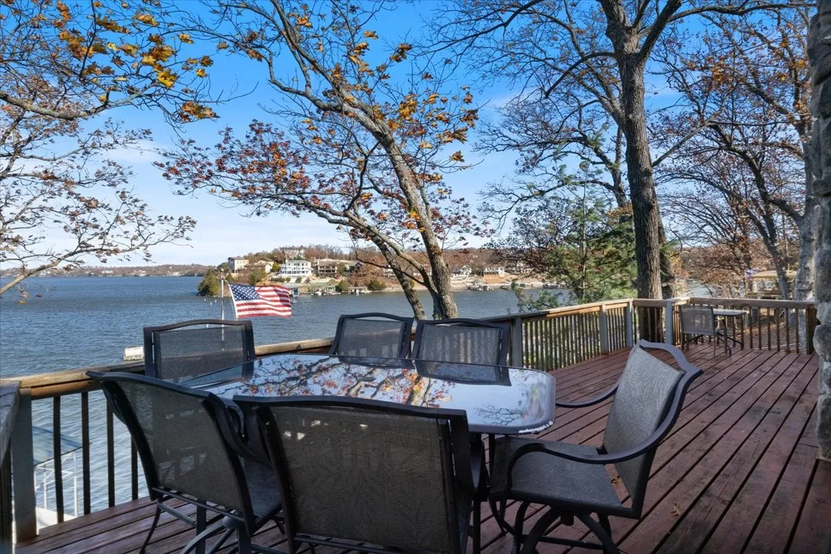 A spacious outdoor deck features a dining table set for a meal, surrounded by trees and overlooking the lake. The American flag waves gently, and the scene offers a clear view of the tranquil water, ideal for alfresco family gatherings and sunset dining.