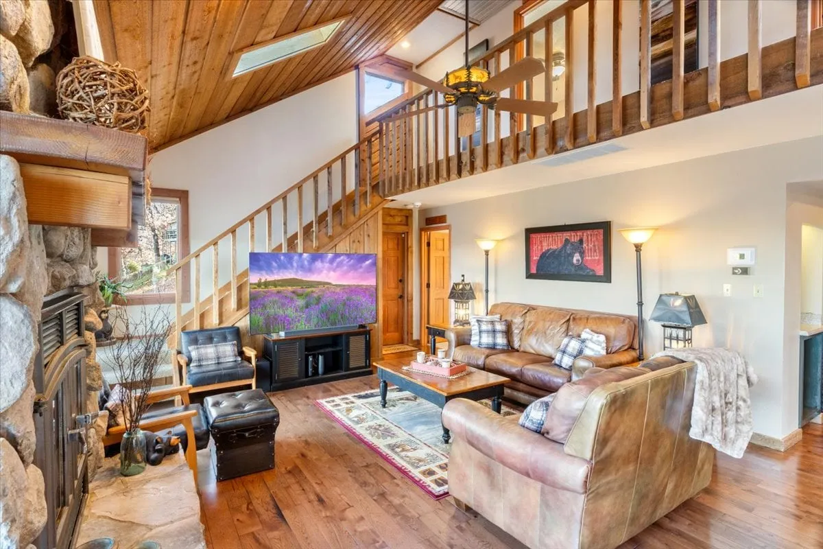 A welcoming living area featuring a handsome stone fireplace, plush sofas arranged for conversation, a large television, and a wooden staircase leading to the upper level. Large windows bring in abundant natural light and offer glimpses of the outdoors, enhancing the cozy yet open feel.