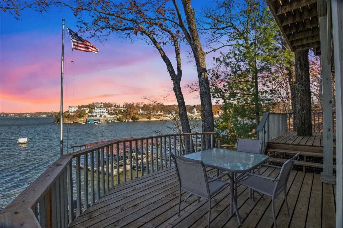The image showcases a spacious deck overlooking a tranquil lake, with comfortable outdoor dining furniture, an American flag waving gently, and a panoramic water view perfect for watching boats pass by. The deck is set up for relaxation and gathering, suggesting ample space for barbecues and sunset dinners, surrounded by nature's beauty.