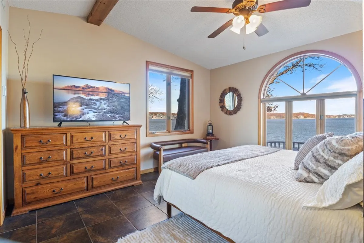 A serene bedroom featuring a comfortable bed, TV for relaxation, and windows overlooking the water. A dressing mirror adds convenience, and the room is bathed in natural light, enhancing its restful atmosphere.