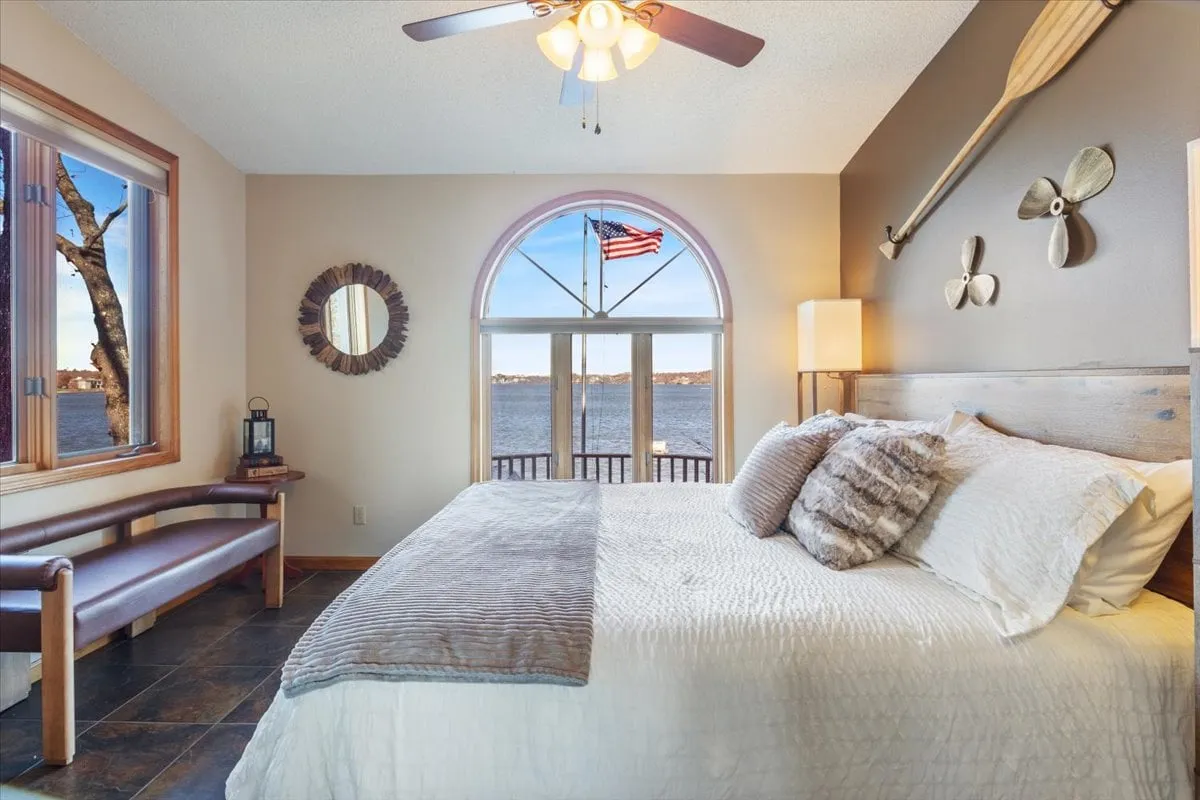 A tranquil king suite with plush bedding, a large window framing an expansive lake view, decorative oars on the wall, and a ceiling fan overhead. The space feels luxurious yet cozy, with soft natural light pouring in and a comfortable bench perfect for relaxing by the window.