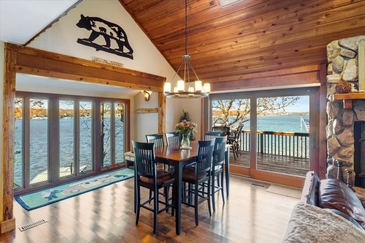 A warm, open dining area with a large wooden table set beside expansive windows offering panoramic lake views. The space features a cozy stone fireplace, rustic wooden beams, and decorative elements like a bear sculpture, all bathed in natural light. The arrangement encourages communal meals and conversation, with the tranquil lake as a constant backdrop.