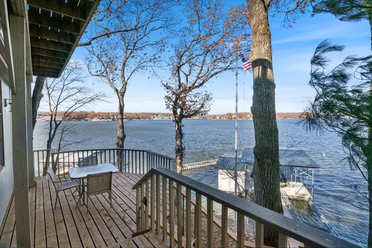 A spacious lakeside deck overlooking the water, surrounded by trees and featuring an American flag. The deck provides ample outdoor seating and stunning sunrise or sunset views across the tranquil lake, creating an ideal spot for group gatherings or quiet reflection.