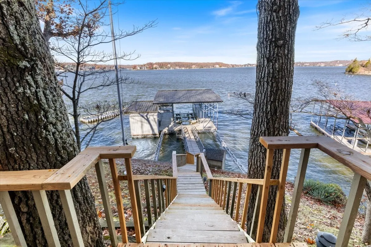 A scenic view looking down wooden stairs toward the water and a dock, framed by greenery. The setting is serene, evoking early mornings or evenings spent fishing or relaxing by the lake.