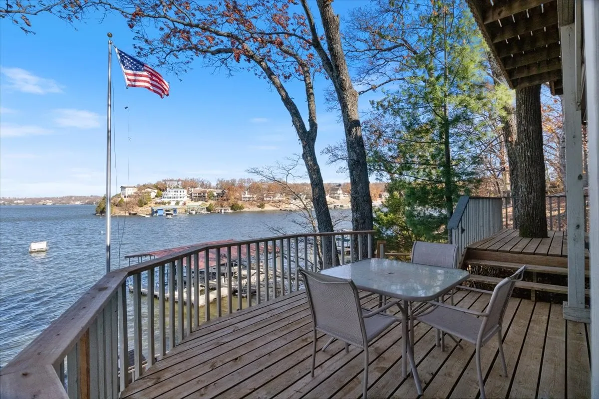 A serene outdoor dining area on a wooden deck, with a table and chairs set against a panoramic lake view, bordered by lush trees and an American flag. The tranquil setting is perfect for outdoor meals, informal meetings, or winding down as a group.
