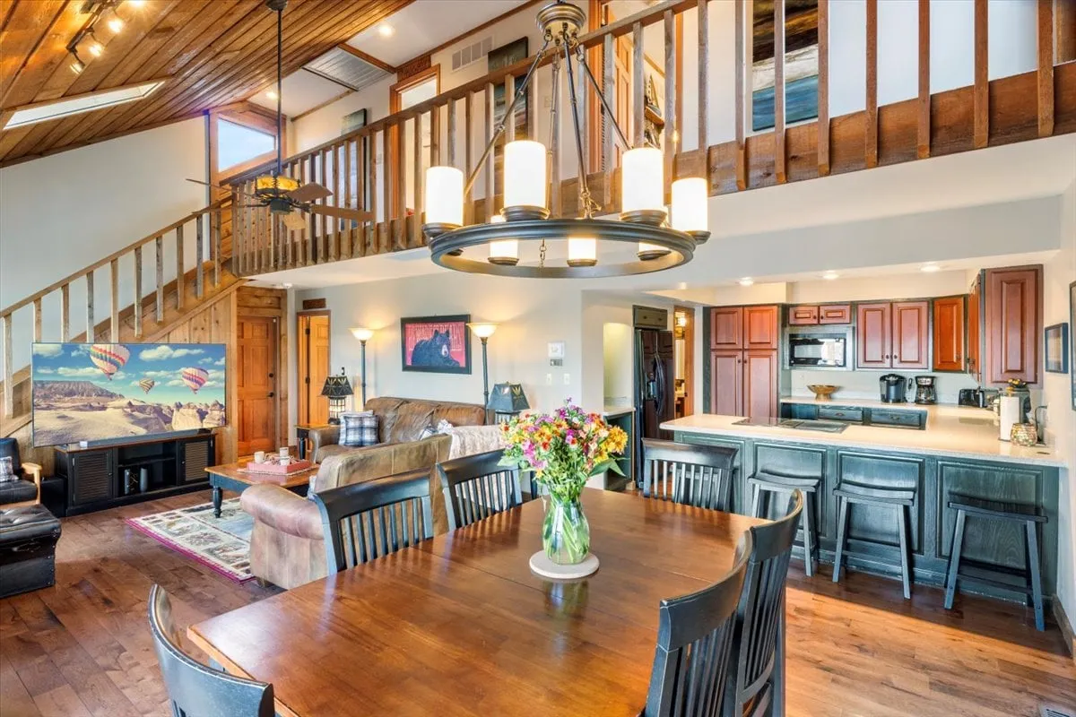 A broad view of an open-concept living and dining space, featuring a large dining table with a floral centerpiece, adjacent kitchen area, and comfortable sofa seating. The room is accented by decorative wall art and benefits from ample natural light, showcasing how the spaces flow together for easy entertaining and communal living.