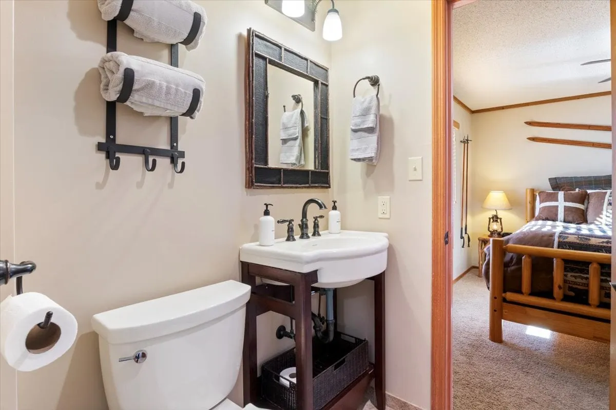 A bright, inviting bathroom with a vanity, large mirror, towel racks, and a glimpse into the adjacent bedroom. The space feels functional yet comfortable—ideal for families or groups needing convenient bathroom access.