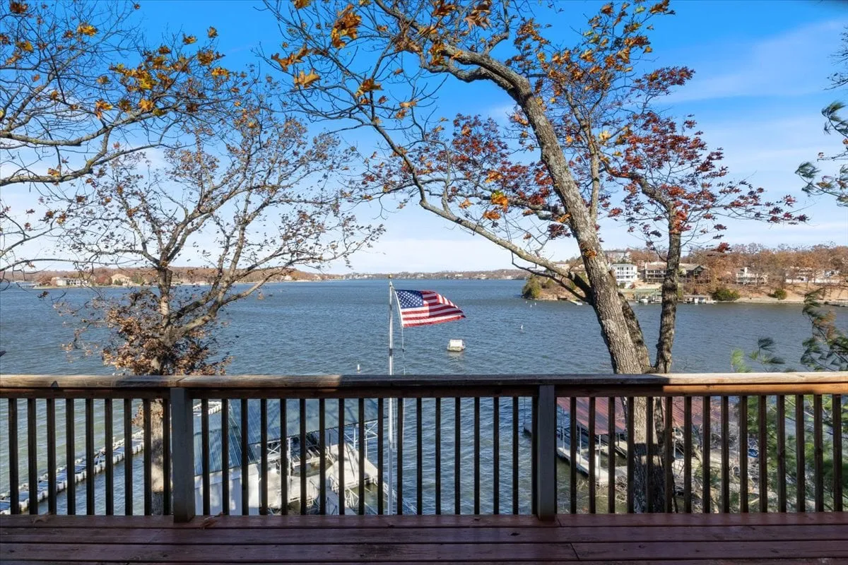 A wide lake view with an American flag, trees lining the shore, and a wooden dock stretching out over the water. The scene is peaceful and inviting, suggesting opportunities for boating, fishing, or simply enjoying nature.