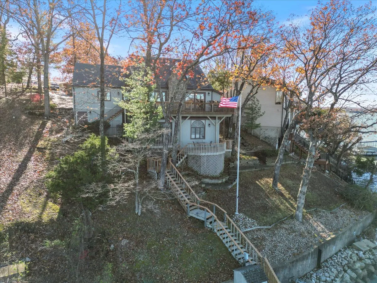 An inviting exterior shot featuring a house with a porch, an American flag, and a staircase leading down to the water. The scene is surrounded by trees with autumn foliage, highlighting the property's connection to both the lake and nearby nature trails.